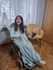 Na fotografiji autorka u pepito dugoj suknji i letnjem džemperu s roza majicom, duge smeđe kose i s naočarima, u kolicima, okrenuta je leđima prozoru s belom zavesom. Pored prozora, malo iza nje s leve strane je žuta fotelja s nogarima u tamnijoj nijansi boje drveta i jastukom na toza, lila i žute štrafte. Vidi se i deo parketa i garderobera u bojji ibrtioce ili svetlijoj boji drveta.